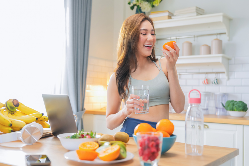 Asian healthy adult woman in sporty casual attire smiles confidently while working from home in daylight, holding a glass of water and fresh strawberries after completing her yoga and stretching