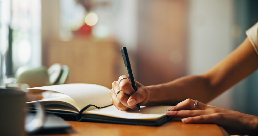 Home, person and hands with notebook for writing, schedule or daily reflection for wellness. Closeup, writer and student with journal for assessment reminder, to do list or thoughts for mental health