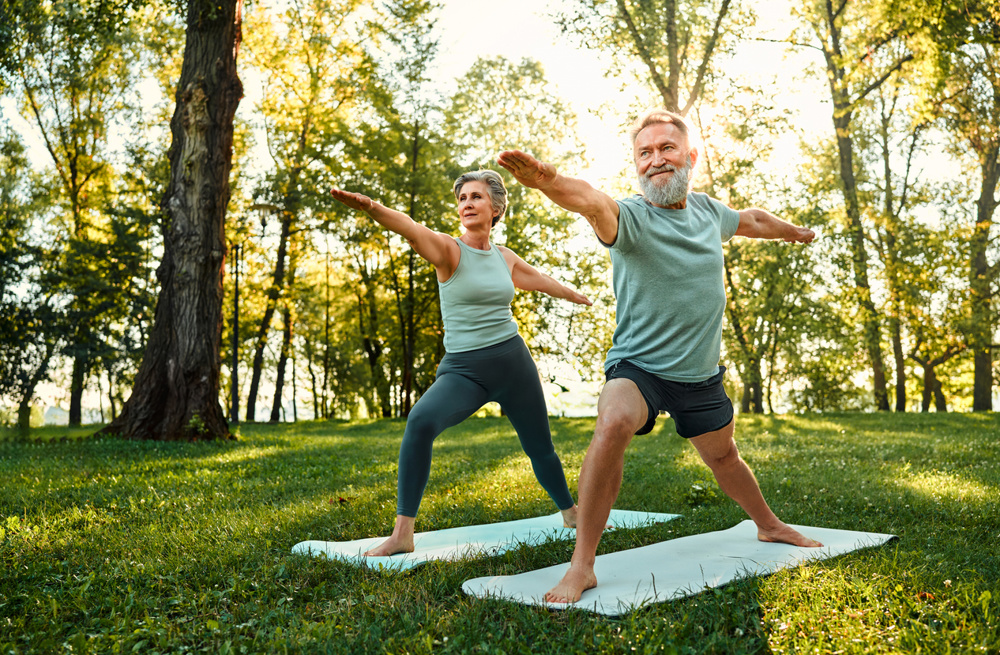Determined family couple standing on rubber mats in national park and performing warrior asana pose. Aged man and woman showing strength and flexibility when practicing yoga